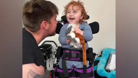 Supplied A little girl standing up in her pediatric walker smiling at her father next to her.