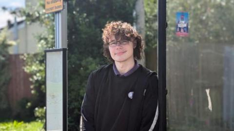 A young man with dark, curly hair and glasses stands at a bus stop. He wears a black top.