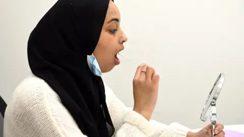 Karwai Tang/Getty Images Woman using a mirror to guide a Covid test swab in her mouth