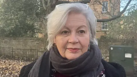Dr Marsha Scott, who has grey hair and white glasses balanced on her head, looking straight at the camera. She is wearing a dark top and a grey scarf.