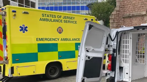 BBC A States of Jersey Ambulance parked up with the doors open at the back. Nearer to the camera is the back of a police van which also has the doors open. 