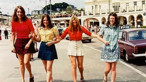 SWNS The same four women link arms on a seaside promenade in 1972. All are wearing bright, distinctive clothes in line with the fashion of the times. A white hotel is in the background.