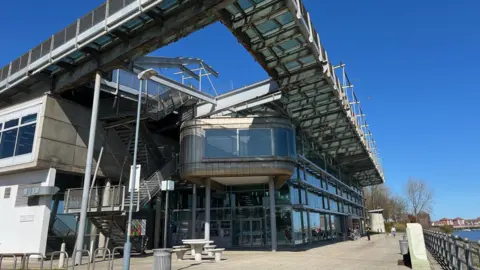 The National Glass Centre is a tall building made of metal panels and glass, overlooking the river Wear. There is seating space and a cfe in front of it, with a dog walker in the distance. 
