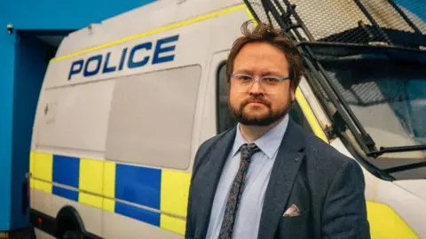 Office of the Cleveland Police and Crime Commissioner Matt Storey standing in front of a police van. He has brown hair and a beard and is wearing glasses, a navy blazer and a light blue shirt with a dark tie.
