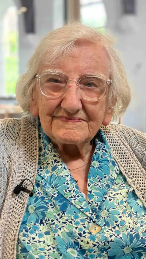 Marjorie, an elderly woman, looks at the camera with a small smile. She has short white hair, and wears clear glasses, a blue floral print shirt and a light cardigan. 