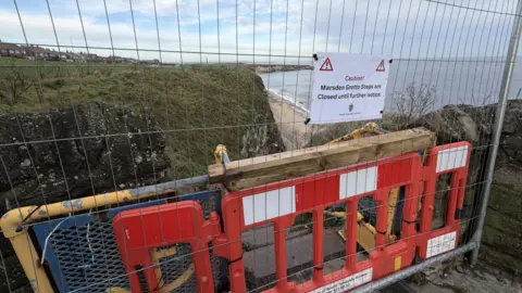 A red plastic and grey metal barrier is placed n front of the cliff steps entrance. A sign reads: "Caution! Marsden Grotto steps are closed until further notice".