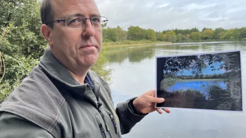 Jonah Fisher/BBC Rob Ballard holding picture in front of water with trees in the background