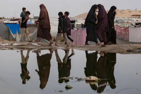 EPA People are reflected as they walk in front of a sewage spill in a makeshift camp on the outskirts of Rafah on 26 April