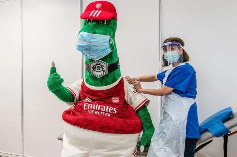 Islington Council Arsenal mascot Gunnersaurus (left) gets his Covid vaccination from an NHS frontline worker