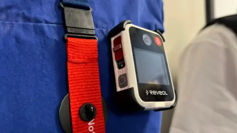 A body-worn camera which has a small screen, clipped to a blue hospital T-shirt 