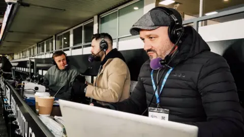 Supplied Ed Dawes wearing a flat cap and coat, commentating on a Derby County match