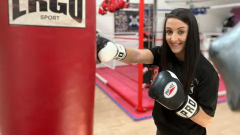 Woman with long black hair wearing black boxing gloves punches a red hanging punch bag, with boxing ring in background.