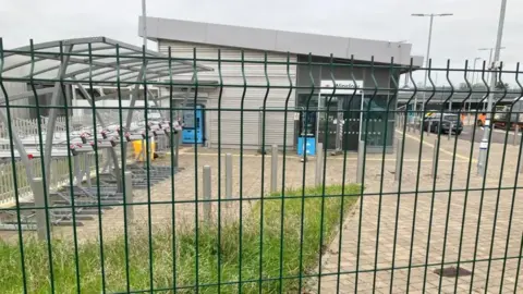 A deserted station building stands behind a wire fence. There are bike racks in the foreground and a blue ticket machine outside the station door. Above the door is a sign saying Winslow.