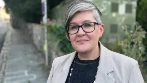 Deputy Haley Camp - A woman withshort coifed grey and black hair, wearing glasses and a grey jacket over a black top.