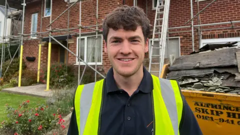 Joseph Gallagher is wearing a yellow high viz vest in front of a brick-built house with scaffolding. Behind him is a yellow skip filled with broken slates and old roofing felt.