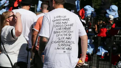 Emma Gibbs A man in a white T-shirt stands in front of a stall selling red and blue bucket hats. Words on the T-shirt read: Listen Up, Shakermaker, Fade Away, Digsy's Dinner, Live Forever, Bring It On Down, Up In The Sky, Slide Away, Cigarettes & Alcohol, Supersonic, I Am The Walrus