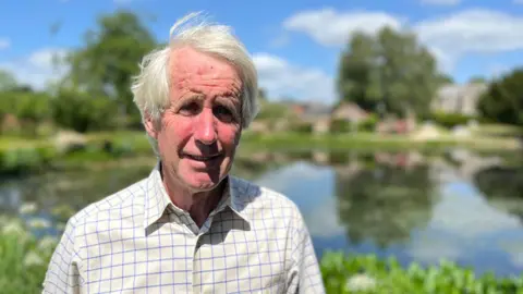 Rupert Thistlethwayte in a check shirt against a blurred background of a large pond and Cadhay House