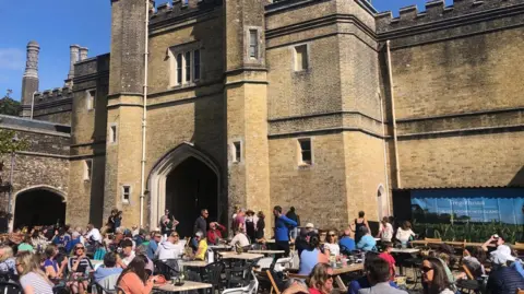 The stable yard at Tregothnan is full of visitors sitting at wooden picnic tables in the sunshine. There is a large historic gatehouse-style building behind them with crenellations and a Gothic -style stone arched entrance.