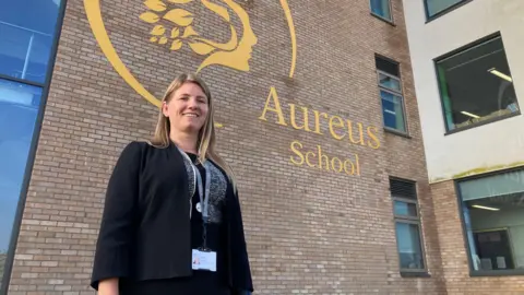 Headteacher of Aureus School, Kirsty Rogers, poses, smiling, in front of the main school building which displays  a large logo of the school reading 'Aureus School Didcot'