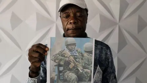 Man wearing white baseball cap poses with a photo of man in army clothing holding a rifle.