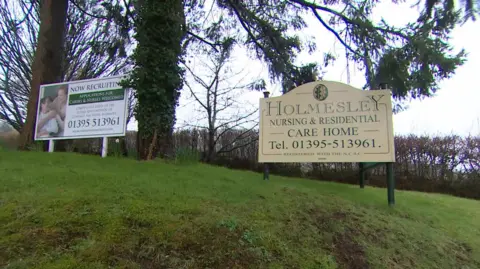 A sign for Holmesley Care Home in Sidford, Devon, next to another sign saying the home is recruiting staff.