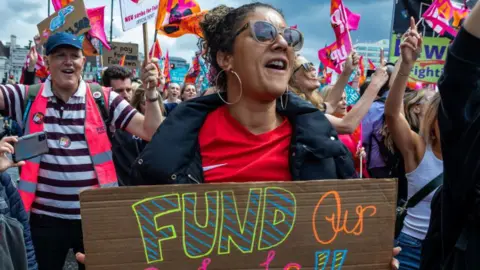 A large group of teachers and education staff on strike are gathered in a street raising their hands and holding flags and placards. In the foreground, a woman with dark hair tied back, hoop earrings and sunglasses, holds a sign that says "fund our schools". She wears a red t-shirt under a black coat. Behind her, a man wearing a blue hat, striped top and a high-vis pink vest holds a flag.
