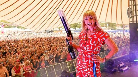 Sara Cox standing with her back to a festival crowd, grinning and holding a confetti cannon