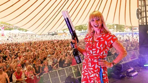 BBC Sara Cox standing with her back to a festival crowd, grinning and holding a confetti cannon