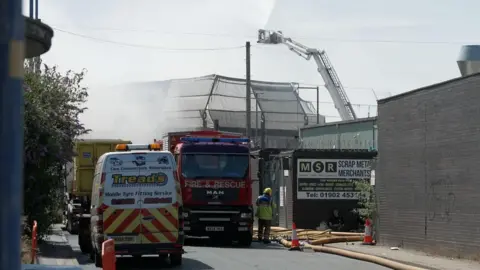 A fire appliance parked on a road in front of an industrial unit, with hoese running along the street. A cherry picker is visible behind. 