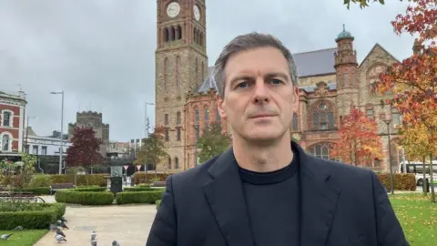 Shows a man in black jumper and black jacket with the Guildhall in Derry in the background
