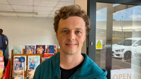 A man in a teal hoodie and black T-shirt with brown curly hair stands in a shop, with greeting cards behind him and a window into a car park to his right.