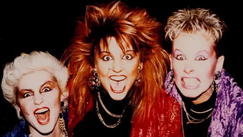 Ruth, Penny and Anita, in a photo from the 1980s, they all have their mouths open, as if shouting. Ruth has white short hair, heavy make up and big earrings, Penny has sticky long hair, a red jacket on, with a black top and large earrings and necklaces on, Anita, has a purple jacket on, purple make up and a black top with necklaces. 