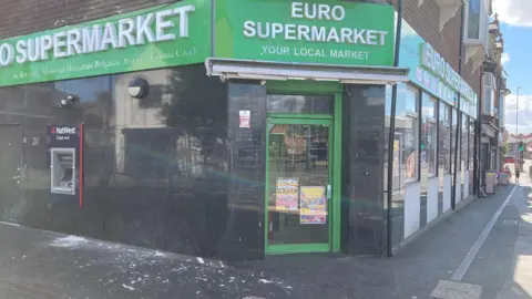 Alex Pope/BBC The outside of a supermarket, with black tiles on it, and paint on the ground. The large sign is green. A road leads down to the right. 