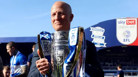 PA Media Birmingham City chairman Tom Wagner with the Sky Bet League One trophy after the Sky Bet League One match at St. Andrew's @ Knighthead Park, Birmingham. He is holding the silver trophy which has blue and white ribbons on it. Behind him is the current stadium and several fans.