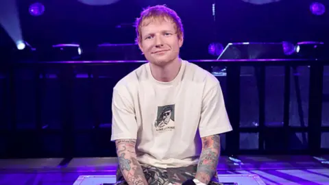 Ed Sheeran wearing a white T-shirt. He's in a room lit by purple light.