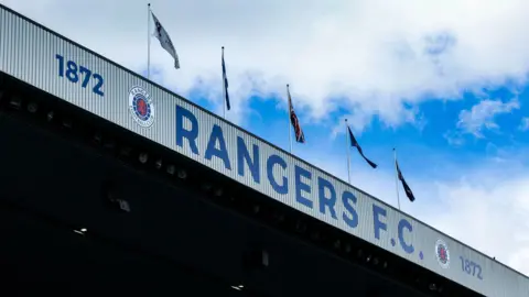 Rangers FC, the club logo and the year 1872 written on part of the Ibrox stadium. There are flags flying above the sign