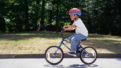 A boy in a white T-shirt and blue jeans with a red helmet on a bicycle, riding on a tarmac path with trees behind him.