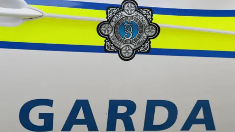 The side of a Garda car door, close up. The garda crest is black and white and the word garda is in block, blue capital letters.