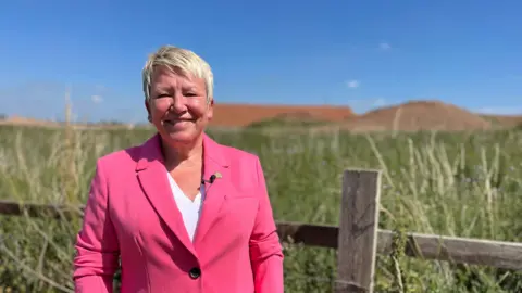 Rachel Taylor, the MP for North Warwickshire and Bedworth wears a bright pink suit with a white top underneath. She has short blonde hair. Behind her is a field and behind that is the High Speed Two works, with two huge piles of earth.