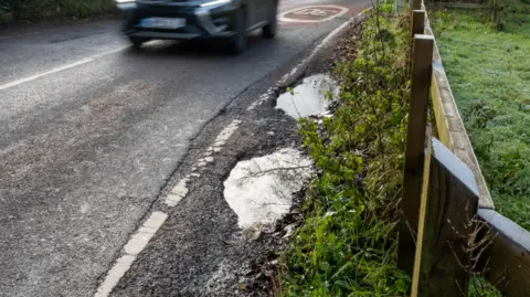 A road travels along a country road. Some rain-filled potholes are on the side of the road next to the grass verge. 