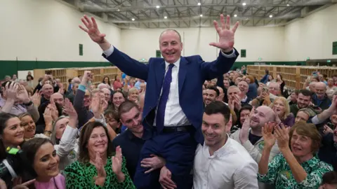 PA Media A man, wearing a navy suit and white shirt, being lifted up by two men. They are surrounded by a crowd of people smiling and clapping. 