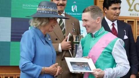 PA Two people stand facing each other during an awards presentation at a racecourse event. One person is wearing a light blue jacket and a wide-brimmed blue hat with decorative ribbon and a feather. The other person is dressed in racing silks featuring turquoise, pink, and white colours and is holding a framed commemorative item. Behind them, several other attendees in formal attire are visible, including one person holding a silver trophy. A large digital display forms the background, showing the word “Juddmonte” and event branding. Bright, even lighting highlights the presentation scene, and the framed item contains text and an image that are too small to read clearly.