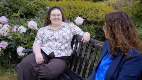 North Horizon Productions A person sits on a wooden bench in a garden filled with blooming pink and white flowers, wearing glasses and a patterned shirt. Another person sits nearby, partially visible, facing toward the first individual as if in conversation. The background is lush with greenery and shrubs, creating a peaceful outdoor setting.