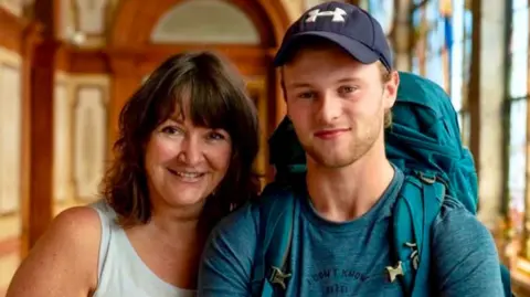 Jo and Sam pose together in an ornate room. He's wearing dark baseball cap and green top and rucksack and smiling, while Jo has shoulder-length brown hair and wears a white vest. She's also smiling.