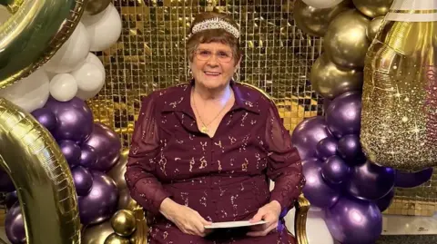 A picture of an elderly lady sat down wearing a purple dress,glasses and a tiara with 90th birthday balloons behind her.