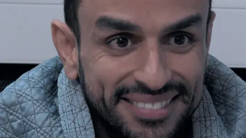 Channel 4 A close up of Mohammed Altaher taken during the filming of 24 Hours in Custody. He has short black hair and a close cropped black beard and is smiling broadly, wearing a padded light blue jacket. 