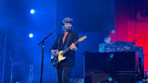 PA Gaz Coombes has a dark beard and medium length dark hair. He wears a flat cap and a dark suit. He is playing a guitar.