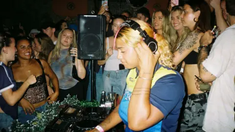 A female DJ adjusts her headphones with her left hand as clubbers dance around her in the background 
