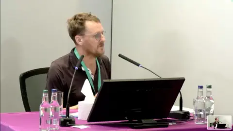 Stuart Ayris looks towards Baroness Lampard. He is wearing a black polo shirt and is sitting in front of a purple table with a laptop in front of him. He is wearing a green lanyard and has bottles of water in front of him. He is sitting in the inquiry room, with a microphone in front of him at Arundel House in central London