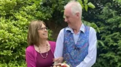 Family photo A woman with bobbed brown hair and glasses, wearing a pink top and cardigan and pink necklace. She is looking at, and has her arm lopped through that of, a man with white hair, wearing a pale blue shirt and patterned blue waistcoat. he is also looking at her. They are stood in front of a large green bush.